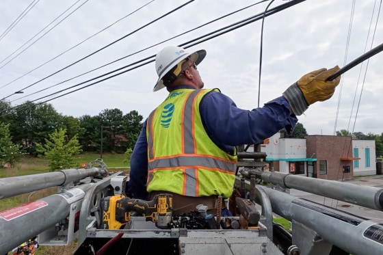 An AT&T technician works on a telephone pole, wearing a light green hazard vest with the AT&T logo and a white hard hat