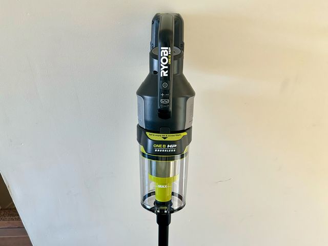 Ryobi 18V One Plus HP Stick Vacuum leaning against a white wall and dark brown floor