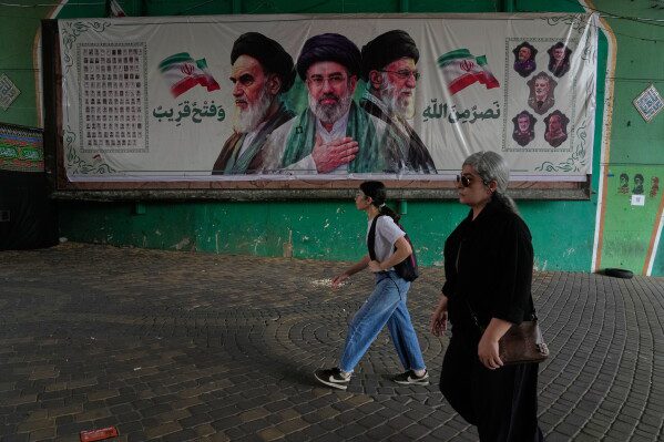 Women walk past a banner showing portraits of the late Iranian revolutionary founder Ayatollah Khomeini, left, Supreme Leader Ayatollah Mojtaba Khamenei, center, and the late Supreme Leader Ayatollah Ali Khamenei, who was killed in the U.S. and Israel strike on Feb. 28, under a bridge in northern Tehran, Iran, Sunday, Aug. 2, 2026. (AP Photo/Vahid Salemi)