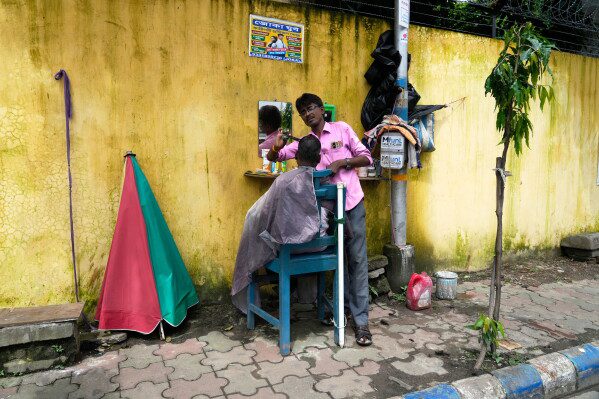 A barber clips a client’s hair at his makeshift open-air shop in Kolkata, India, Saturday, Aug. 8, 2026. (AP Photo/Bikas Das)